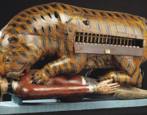 A wooden anamatronic tiger crouches on top of a wooden body of a man dressed in European dress, the tiger is mauling his neck.