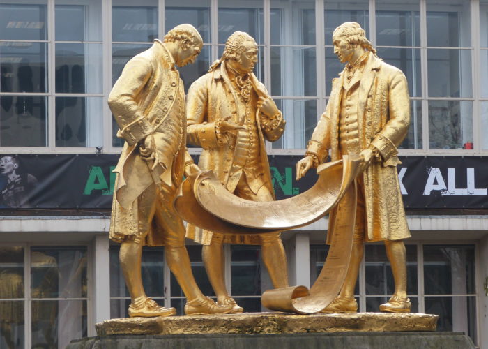 A photograph of a gold statue showing three men dressed in typical Victorian fashion discussing the plans for the railway