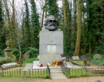 A photograph of the grave of Karl Marx. A large stone plinth is surrounded by flowers, on top of the plinth is a lerge, black statue of the head of Karl Marx.