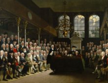 A painting showing the inside of the house of commons, the room is full of well dressed men with white hair. In the centre of the room is a table and a man can be seen speaking to the room.