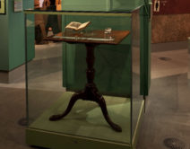 A photograph of the inside of the museum. A small wooden writing desk can be seen in a glass case.