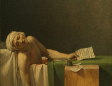 A painting of a dying man sprawled over the edge of his bathtub. He wears a white turban on his head and is clutching a quill in one hand an a piece of parchment in the other. Next to him is a wooden block which reads 'A MARAT. DAVID' at the bottom.