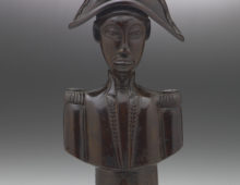 A wooden, sculpted bust of Jean-Jaques Dessaline, he is dressed in military uniform with a feathered, bicorne hat. His face is quite narrow and stylised.