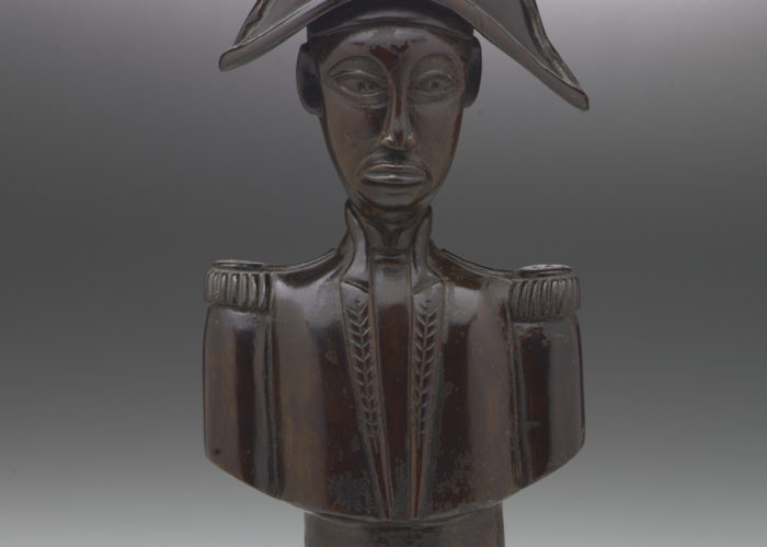A wooden, sculpted bust of Jean-Jaques Dessaline, he is dressed in military uniform with a feathered, bicorne hat. His face is quite narrow and stylised.