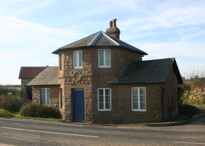 A modern photograph of a tollhouse building on the side of the road. The building is made of stone and has a blue door and white windows