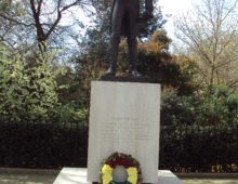 A photograph of a statue in front of a park. The statue is dark brown and shows a man dressed in a formal military uniform. The statue stands on a stone plinth with a wreath of flowers at the bottom. The plinth is engraved with the word 'BOLIVAR'