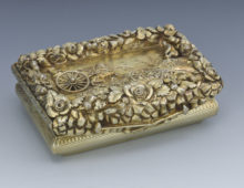 A photograph of a small silver gilt snuffbox with a representation of a coach in the centre of the lid