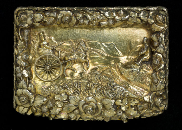 A photograph taken from above of a small silver gilt snuffbox with a representation of a coach in the centre of the lid