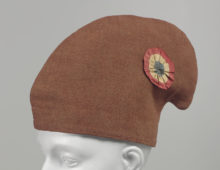 A photograph of a dark orange cap on a manequin head. The cap is hooked at the top and is decorated with a circular, red, white and blue pin.