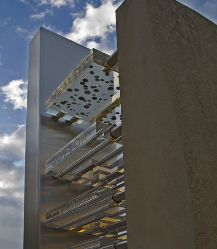 A photograph of the statue taken from below. The photo shows four tiers of clear perspex suspended between two tall towers, one concrete and one metal. A blue and cloudy sky can be seen in the background.