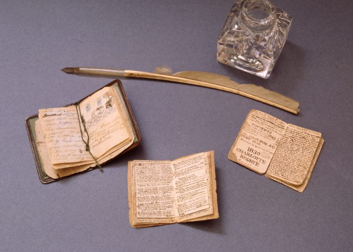A photograph taken from above of three little hand written books, each laying open. Next to the books lies a feathered quill and a crystal glass pot for ink. The books are a similar size to the ink pot.