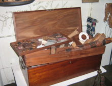 A photograph of a wooden chest with its contents laid out on display. A number of striped fabrics in different colours are draped over the box as well as a beaded necklace. In the foreground a number of trinkets and objects can be seen.