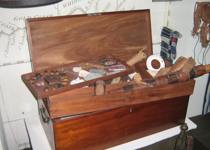 A photograph of a wooden chest with its contents laid out on display. A number of striped fabrics in different colours are draped over the box as well as a beaded necklace. In the foreground a number of trinkets and objects can be seen.