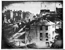 A black and white grainy photograph of a street scene taken from a height. In the bottom left corner a person can be seen having their shoes shined.