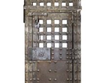 A photograph of a door against a white background. The door is covered in iron and has a large bolt at the top and bottom of the door as well as a big lock in the middle. The upper sections of the door are grated to allow prisoners to see out. The door is rusting in places and looks old.