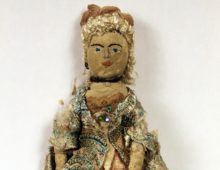 A photograph of a female doll against a white background. The doll is made of textliles and looks rather old and worn. The remains of a courtly outfit can be seen and the doll is wearing a patterned long jacket. The face and hair are that of a woman.