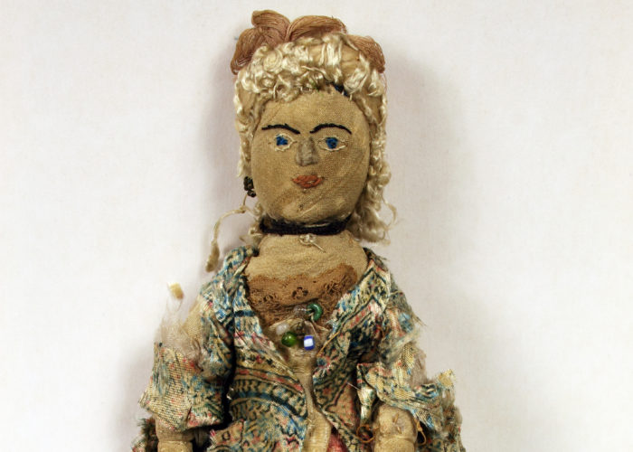 A photograph of a female doll against a white background. The doll is made of textliles and looks rather old and worn. The remains of a courtly outfit can be seen and the doll is wearing a patterned long jacket. The face and hair are that of a woman.