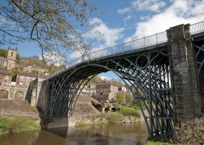 A photograph of a green iron bridge over a river with a small town in the background